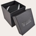 A black necktie with white diagonal stripes, presented inside an open black gift box with its lid leaning against it. The lid is embossed with the Northumbria University Newcastle crest and text.