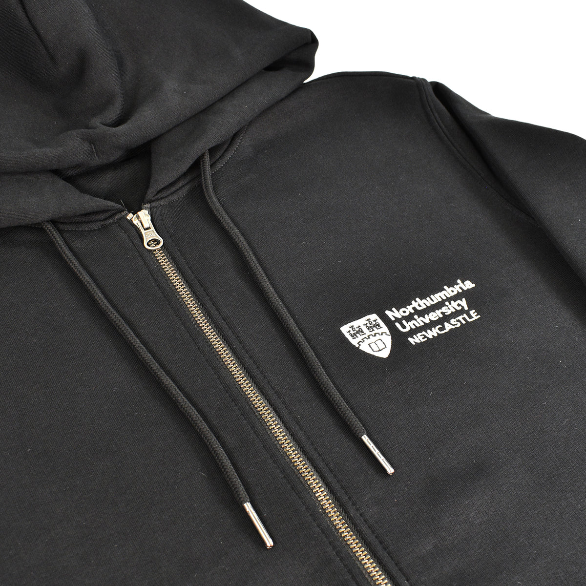 A close-up of a black zip-up hoodie with a white embroidered logo for Northumbria University on the chest. The hoodie features black drawstrings with silver metal tips and a gold-toned zipper.